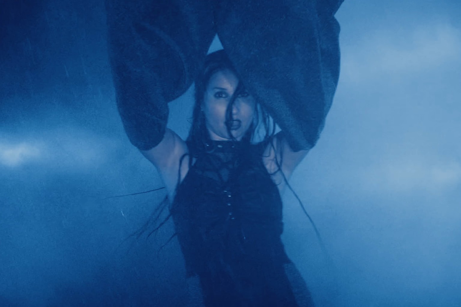 Fotograma del videoclip de Tormenta de Xeria: Marina, con los brazos en alto y mangas azules, bajo la lluvia y entre nubes en tonos azules